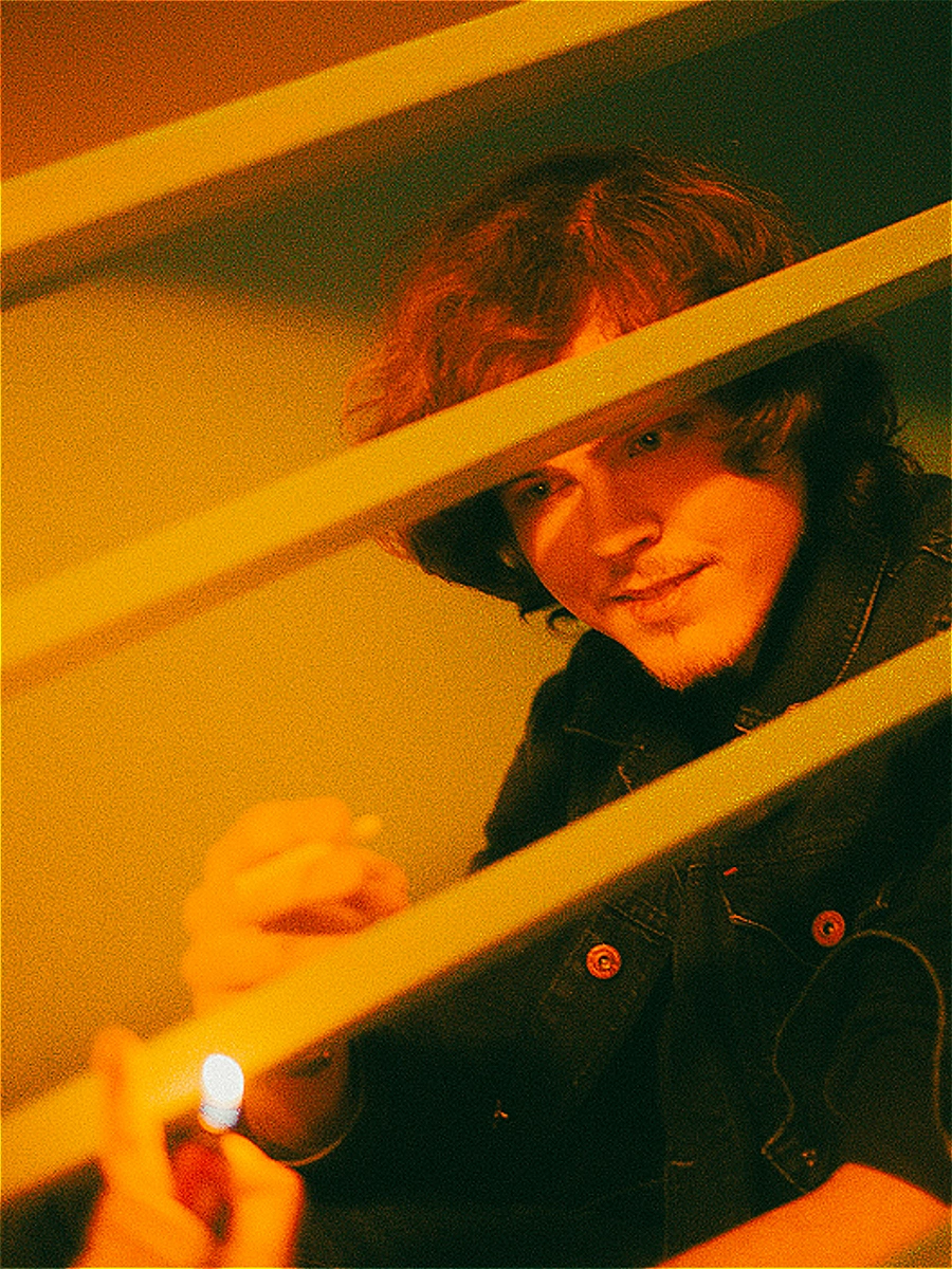 Jeune homme aux cheveux bouclés en veste en jean derrière les barreaux d'un escalier en bois, éclairé par une lumière ambrée et une étincelle de briquet. Photo par Interviolate.