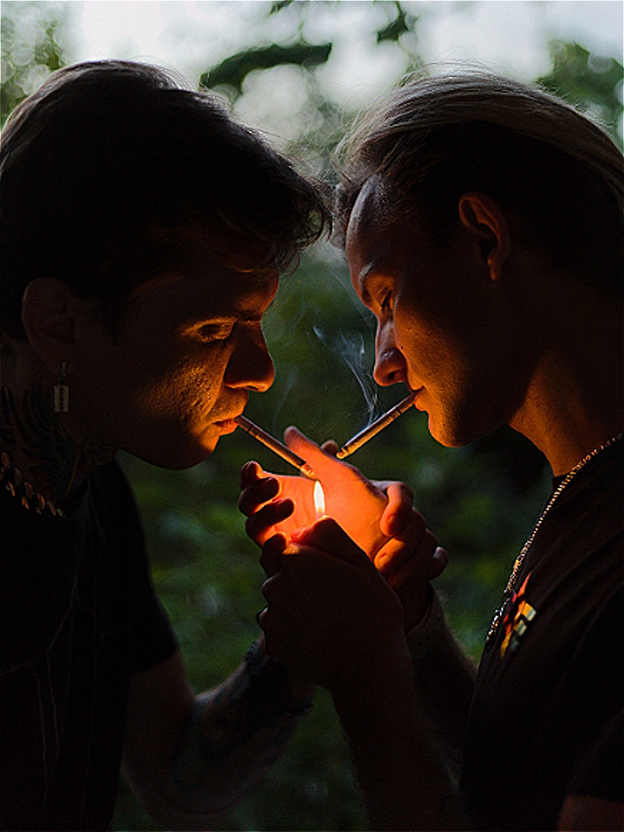 Deux jeunes hommes dans l'obscurité se penchant l'un vers l'autre pour allumer des cigarettes à la flamme d'un seul briquet. Photographie d'art par Interviolate.
