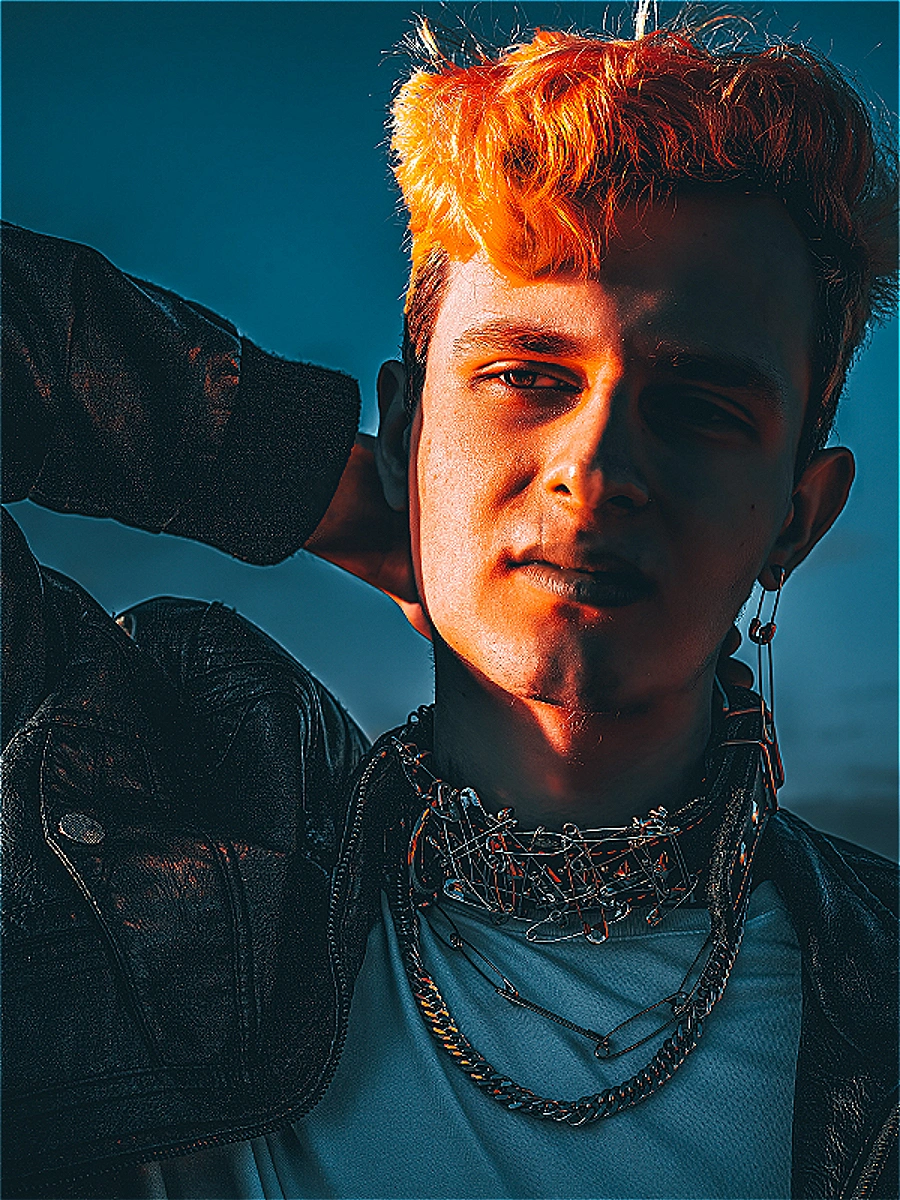 Jeune homme aux cheveux orange néon éclatants, veste en cuir noir et ras-du-cou en épingles de sûreté sous éclairage clair-obscur dramatique. Photographie par Interviolate, tous droits réservés.